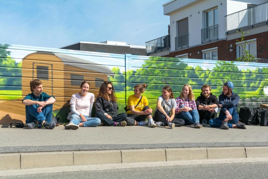Hier sind 8 Mitglieder des klimaentscheids zu sehen, die in einer Reihe vor einer Lärmschutzwand sitzen und sich unterhalten. Die Lärmschutzwand ist mit Graffiti besprüht und es ist eine Bauhütte mit Wald im Hintergrund zu sehen.